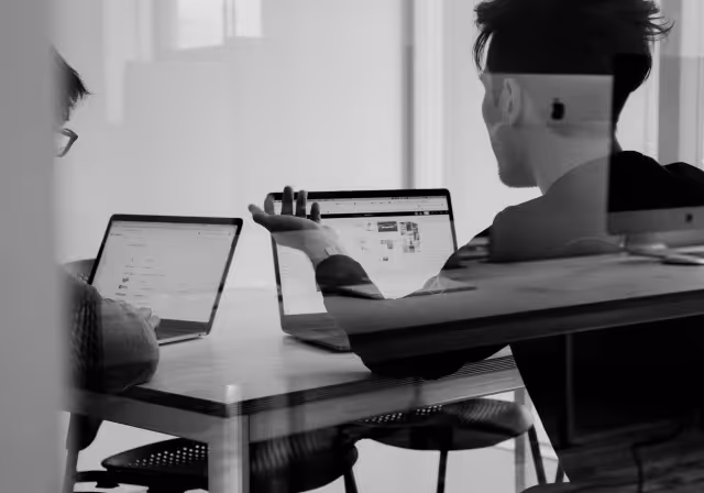 Two people seated at a table with laptops, one gesturing with hand as they discuss work in a glass-walled office.