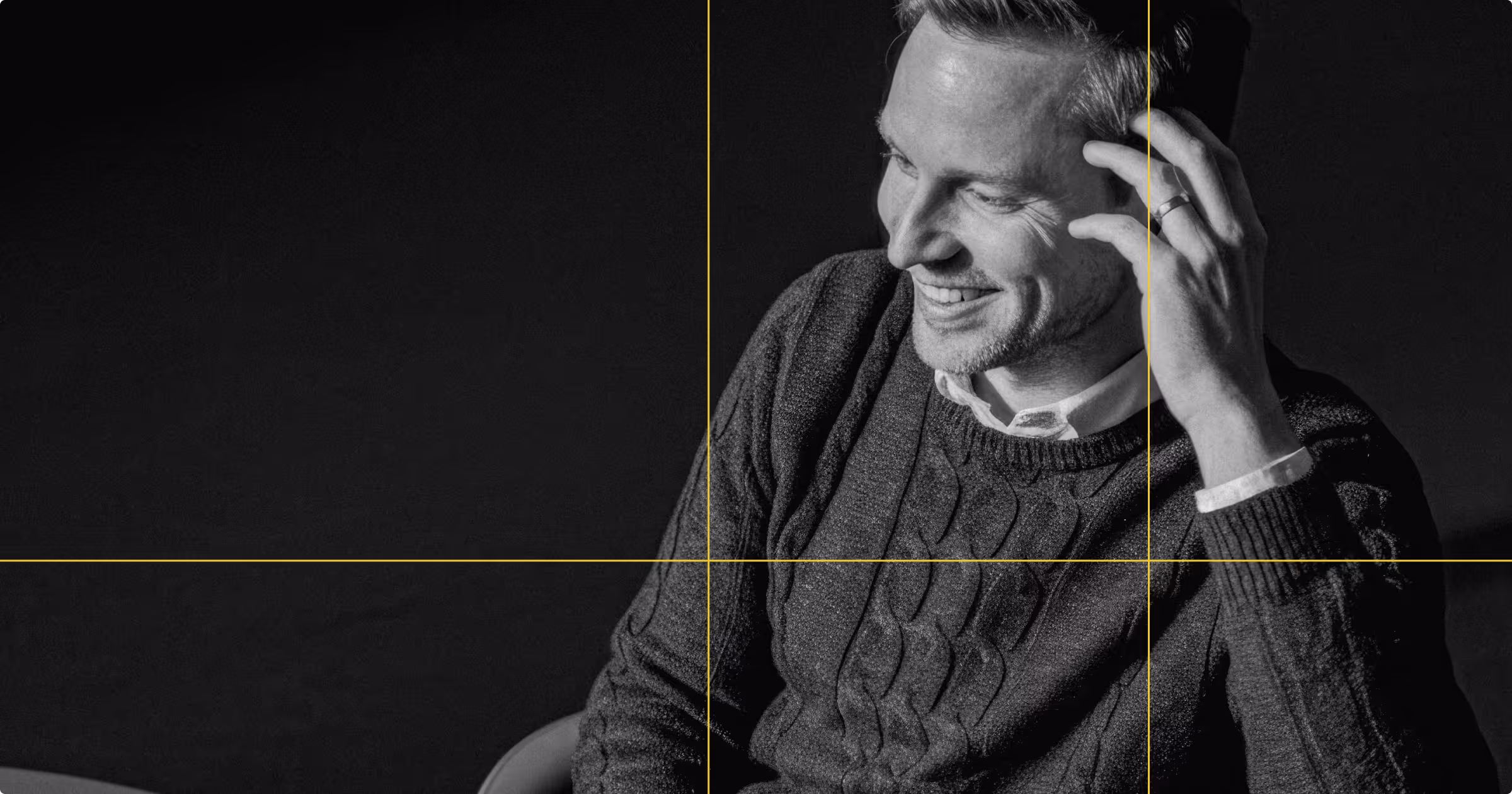 Smiling man in a cable-knit sweater touching his hair against a dark background.