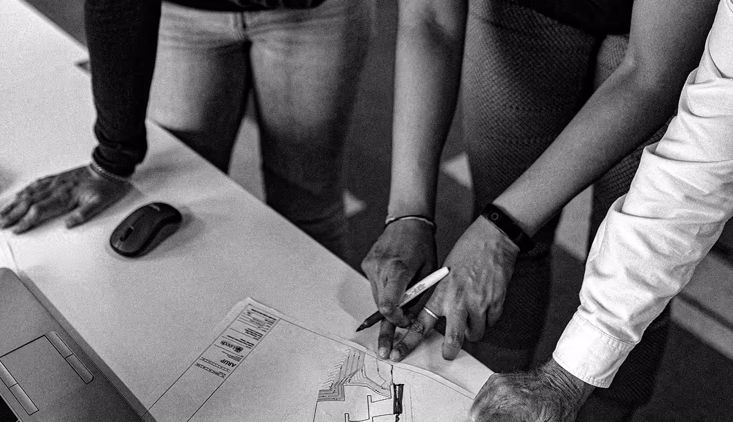 Three people collaborate over architectural plans on a table, one holding a pen and pointing.