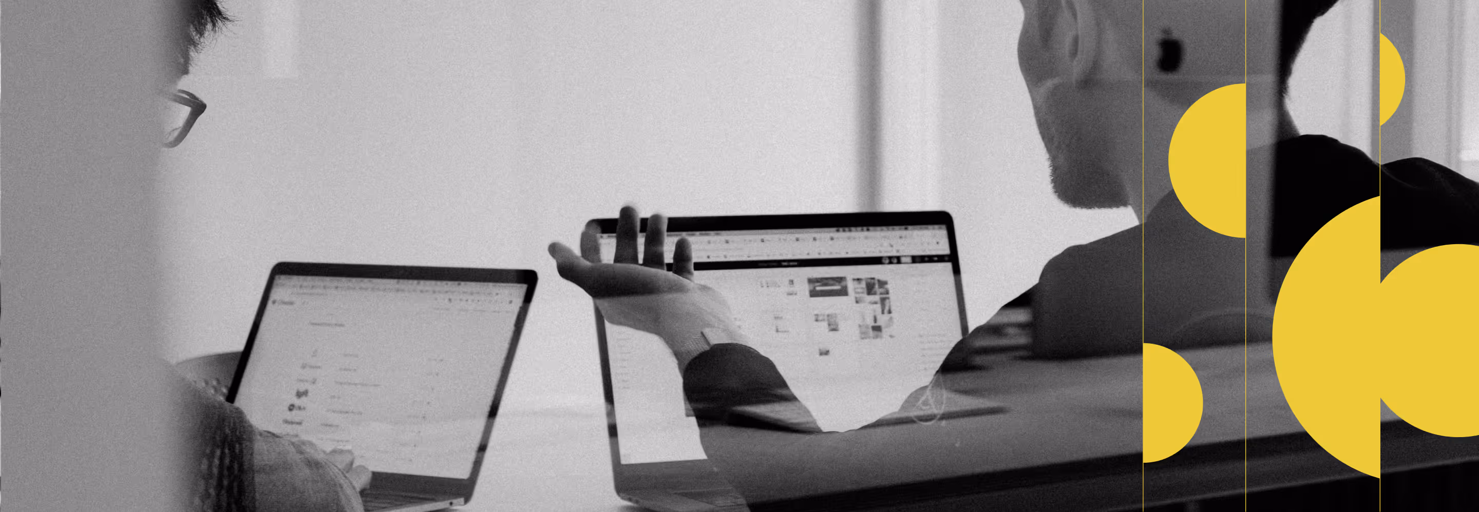 Two people working on laptops, one gesturing, seen through a glass window with yellow design shapes.