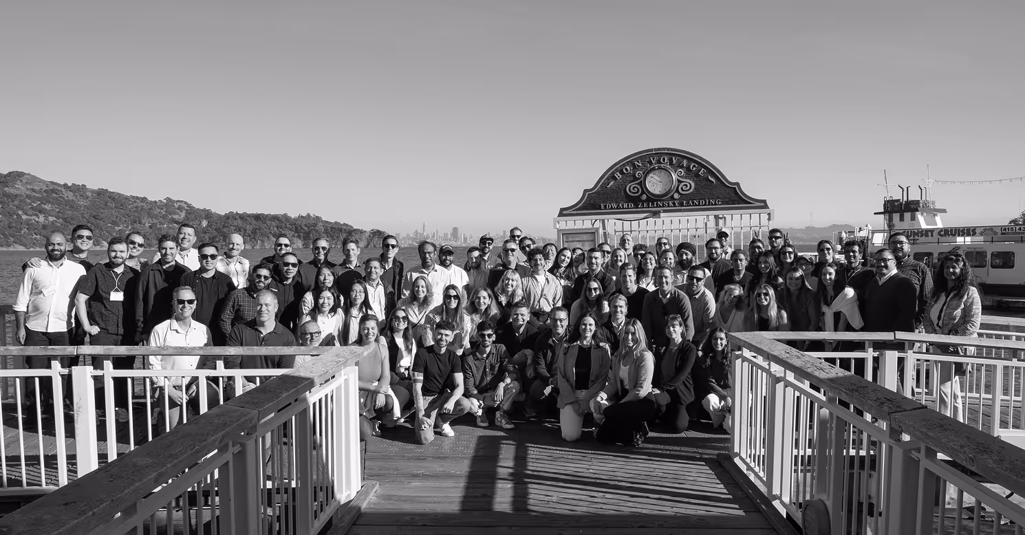 Large group of people posing on a wooden pier under a sign reading 'Horn Voyage Edward Zelinsky Landing' with a body of water and a distant city skyline in the background.