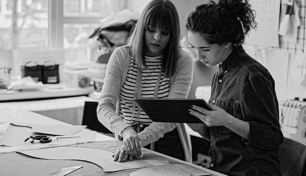 Two women working on sewing patterns at a table, one using a tablet and the other pointing at fabric.