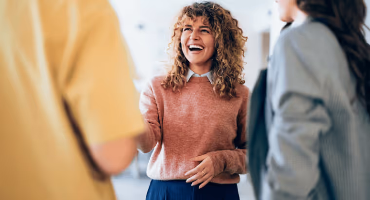 Curly-haired woman in a pink sweater smiling and talking with two people indoors.