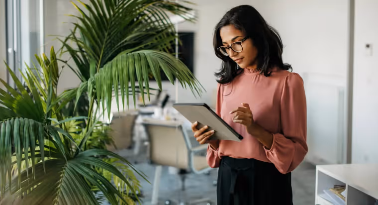 Woman in glasses and pink blouse using a tablet in a modern office with large green plants.